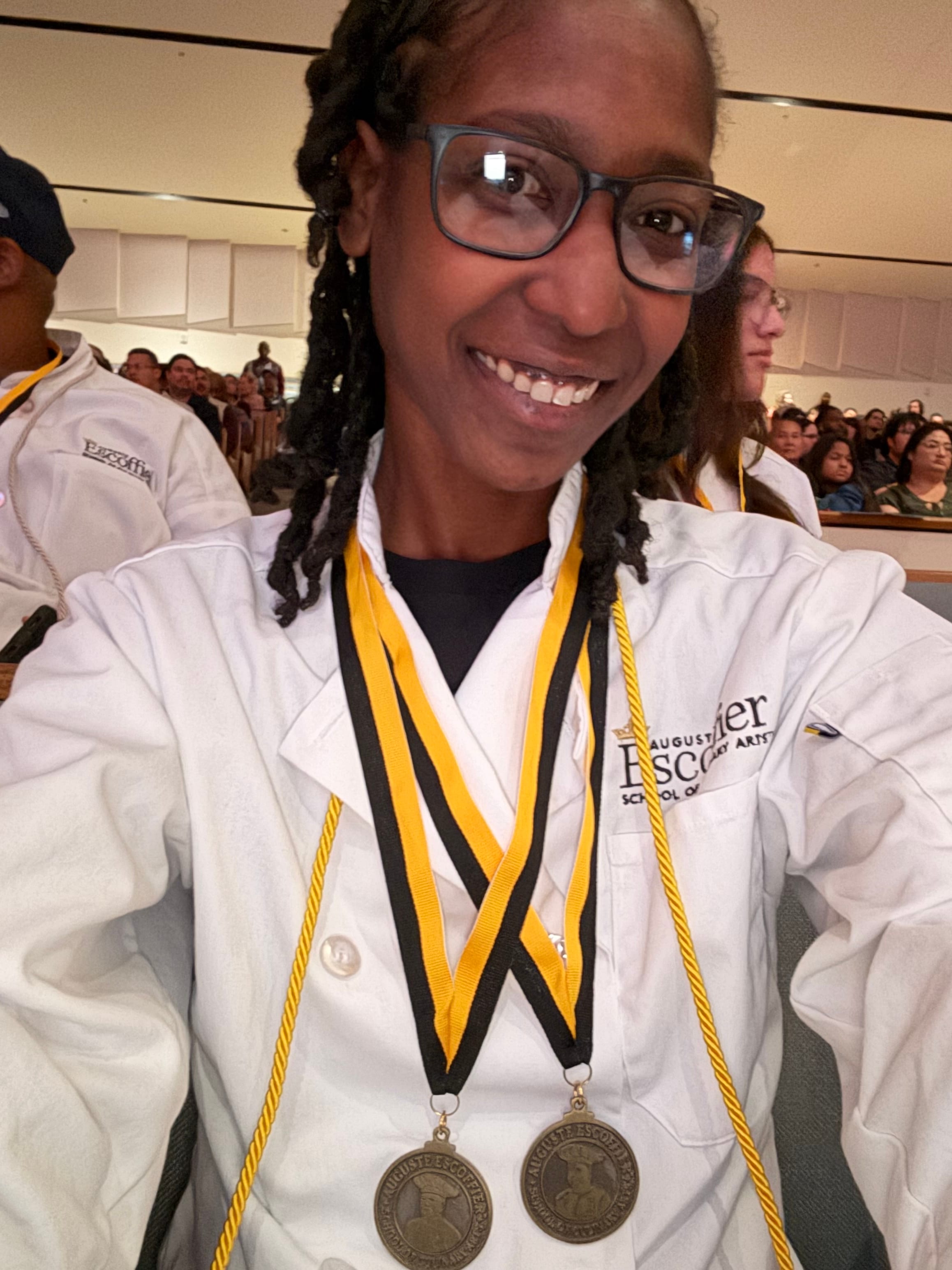 Maya in her chef coat, smiling after her Escoffier culinary school graduation